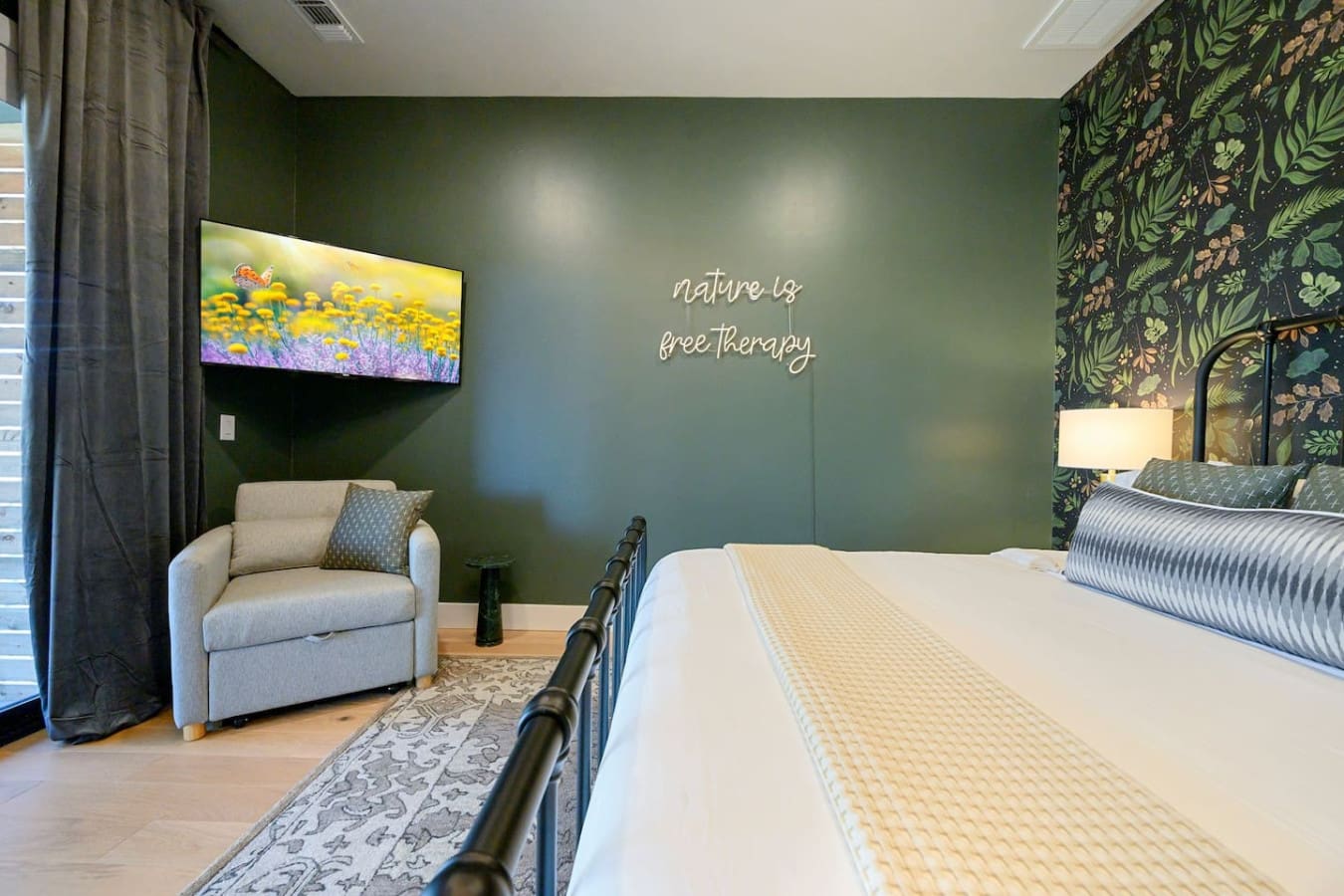 This serene bedroom strikes the perfect balance between moody and cozy with its soft sage-green walls, neon "nature is free therapy" sign, and that inviting reading chair tucked by the window—basically your own personal wellness retreat without ever leavi