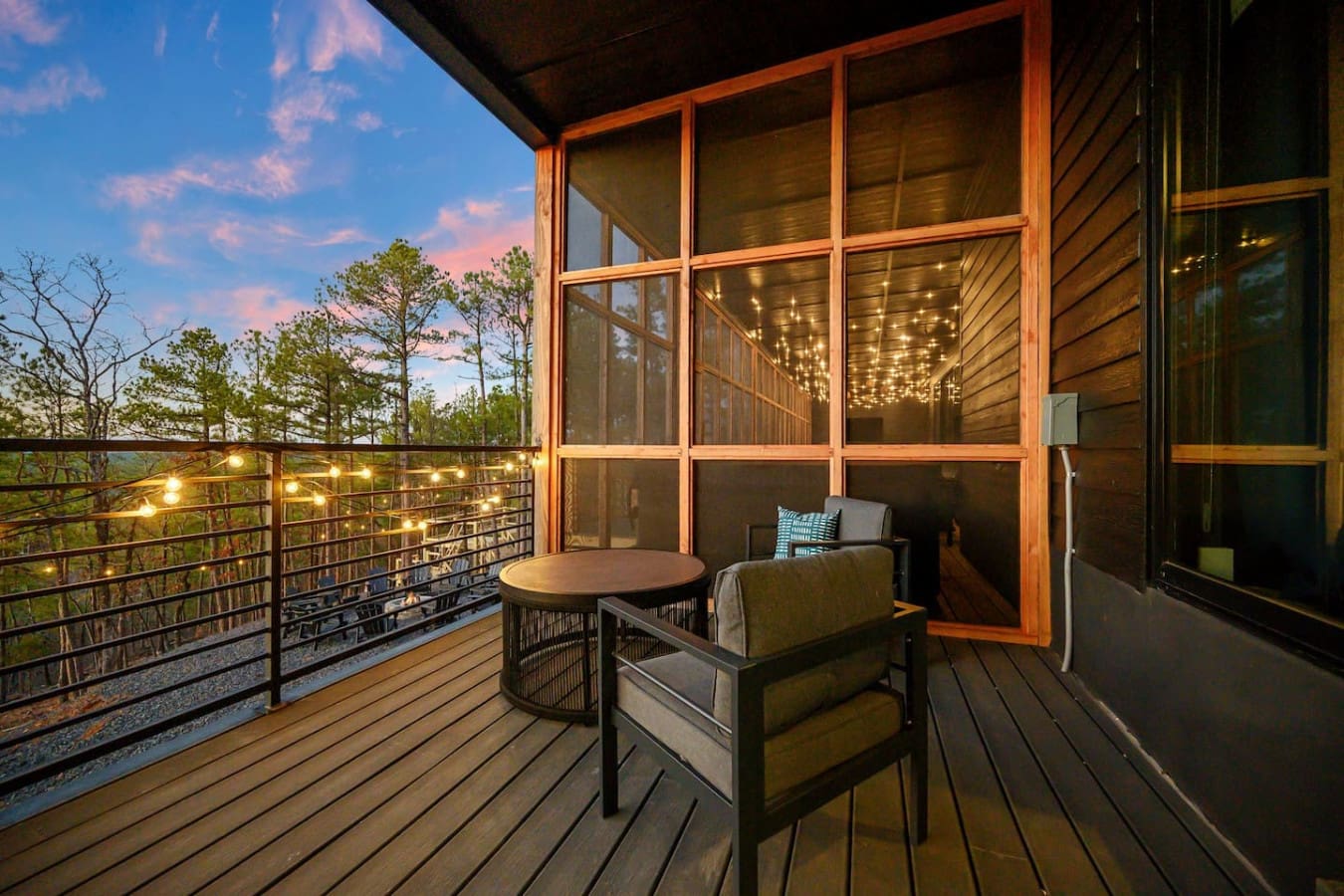This deck basically feels like your own private treetop retreat—cozy seating tucked under those glowing string lights, massive windows framing the glowing interior behind you, and an endless forest view that just keeps getting better as the sun sets.