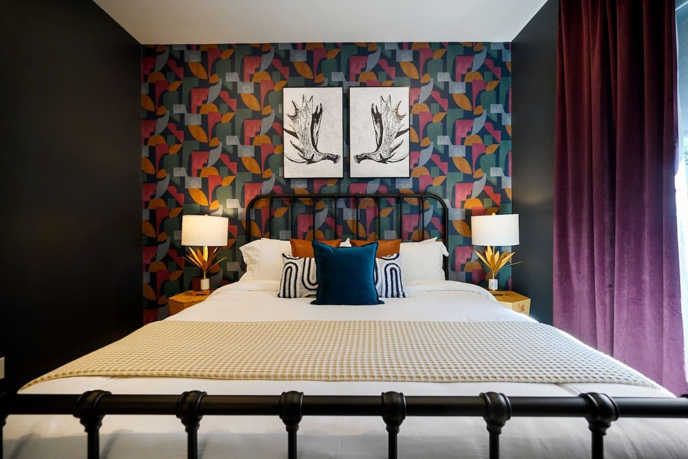 This bedroom is basically a bold design statement with that eye-catching patterned wallpaper, sleek black bed frame, those amazing antler art prints above, and rich jewel-toned curtains that somehow make the whole room feel both artistic and incredibly co