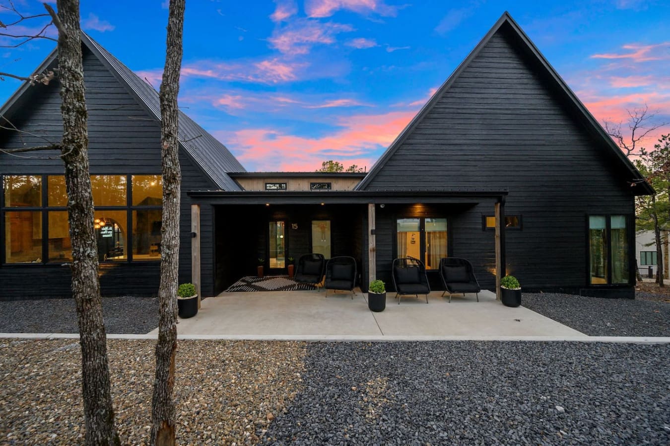 This sleek modern retreat with its dramatic peaked roofs and expansive covered patio basically looks like the perfect spot to unwind with that stunning twilight sky as your backdrop—black siding, warm glowing interiors, and those comfortable chairs all se