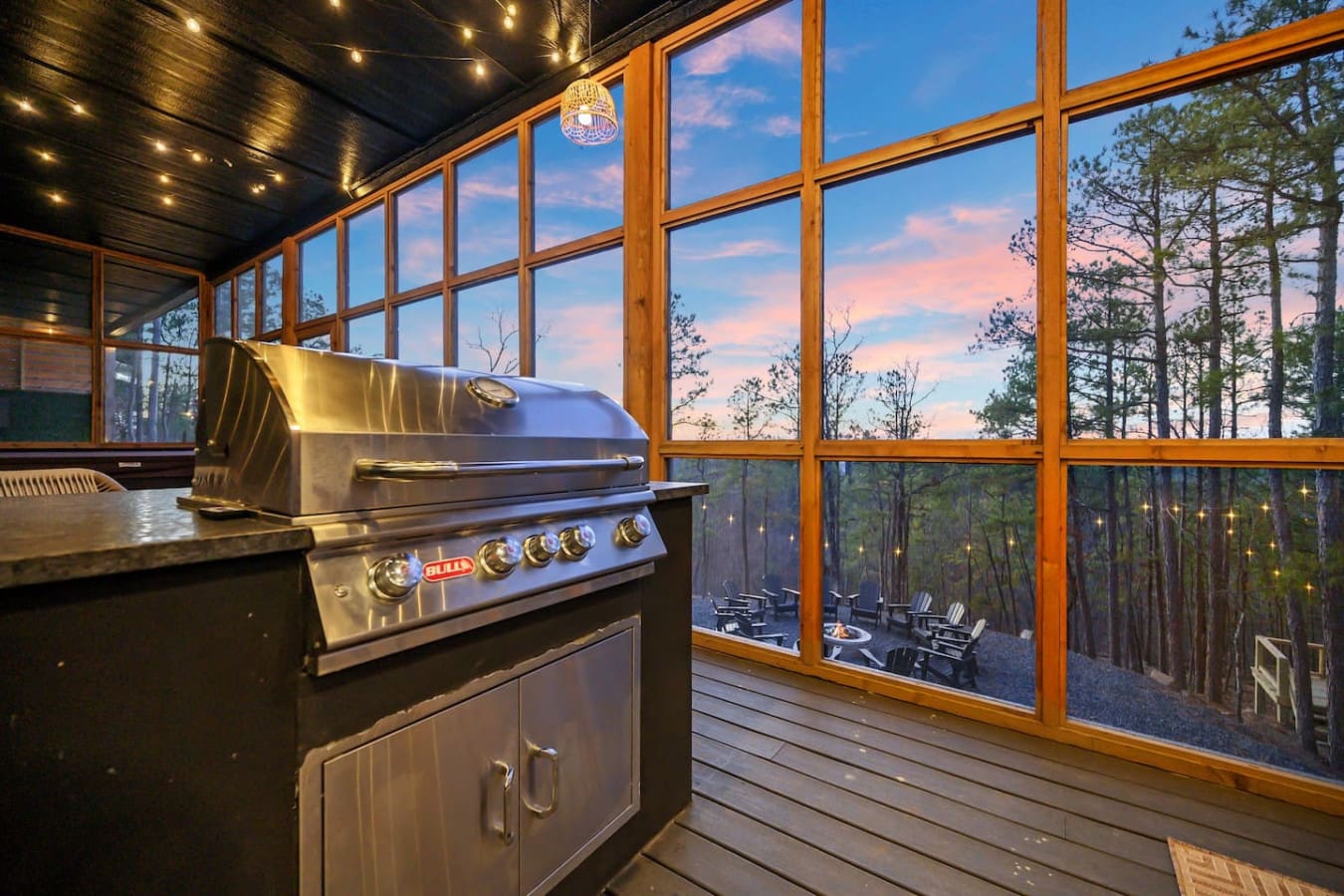 This covered outdoor kitchen with its sleek stainless steel grill, those massive windows framing the sunset sky, and string lights twinkling overhead is basically the dream setup for cooking dinner while watching the forest glow golden at dusk.