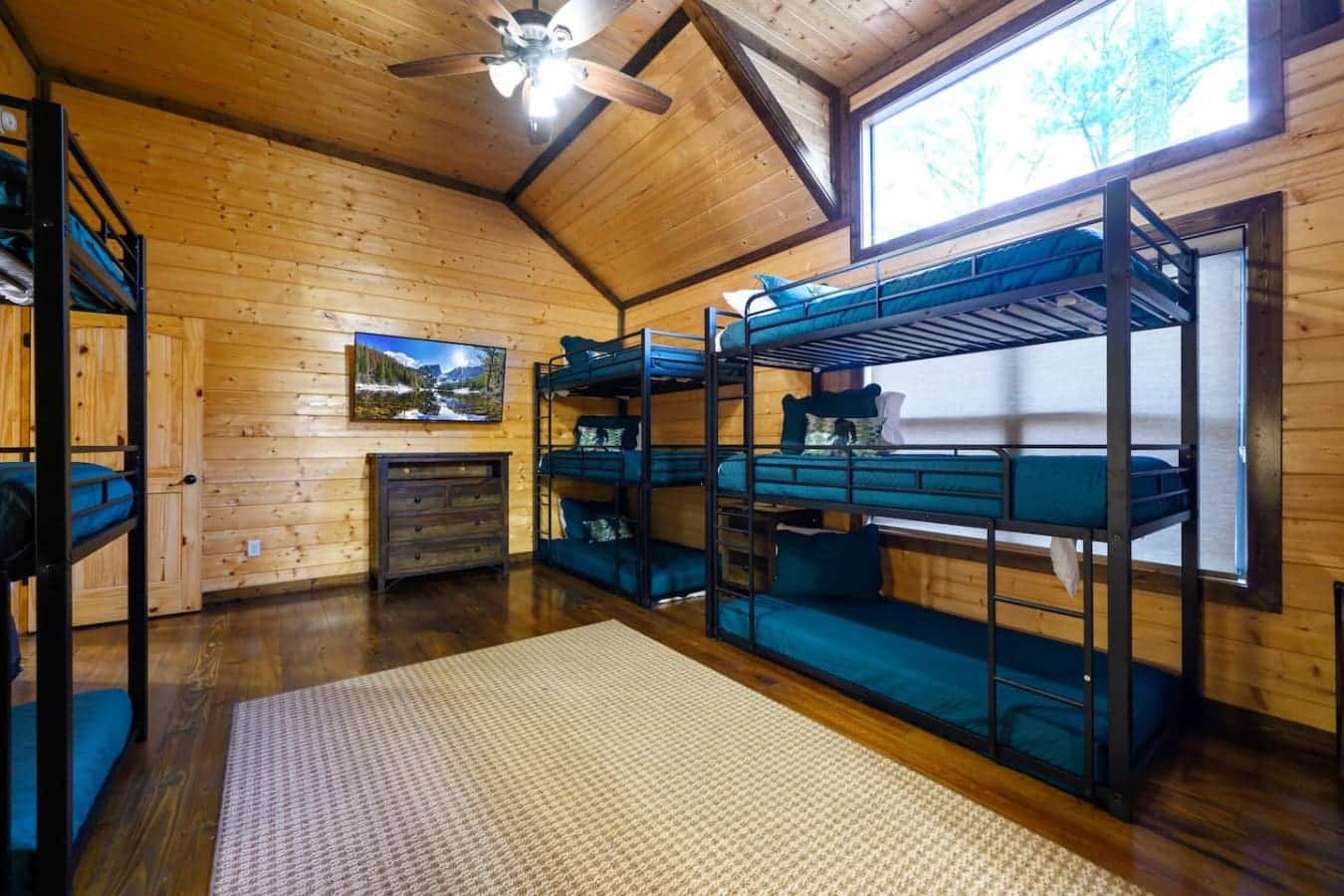 This spacious bunk room sleeps a ton of people without feeling cramped, with multiple bunk beds spread across a huge wood floor, a TV on the wall so there's actually something to do before bed, and enough windows and ceiling height that it feels way more