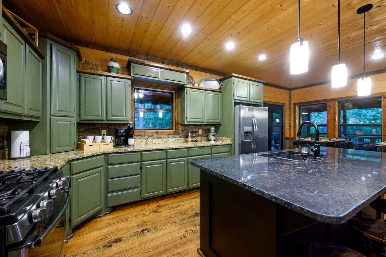 This kitchen basically has everything you'd actually want to cook with—the green cabinets are a vibe, the island's massive, and those windows looking out at the trees make chopping vegetables way less of a chore.
