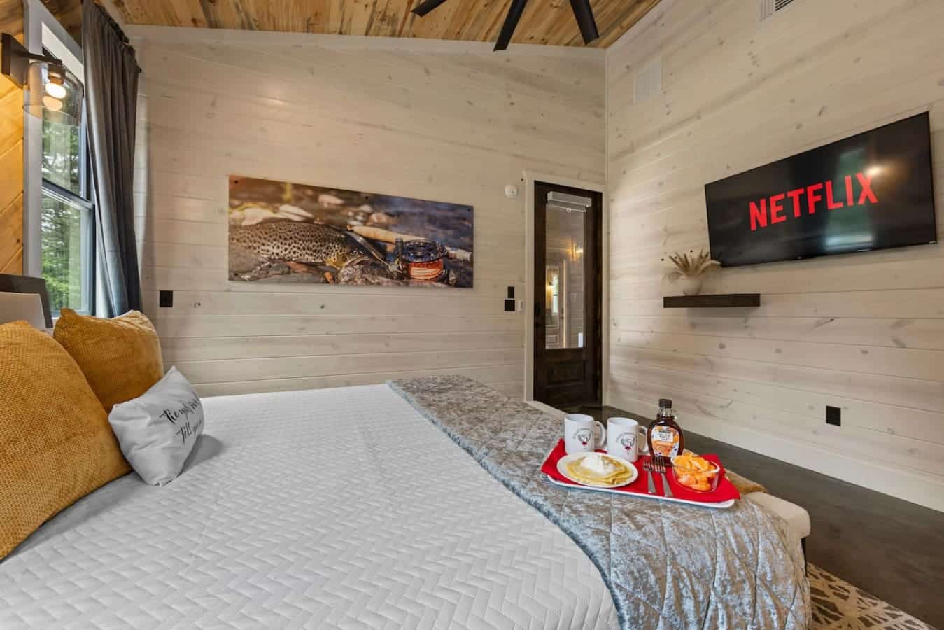 This view highlights the perfect lazy morning setup: cozy pillows, breakfast in bed, and Netflix ready to stream — all set in a beautifully rustic, wood-paneled bedroom that oozes comfort and style.