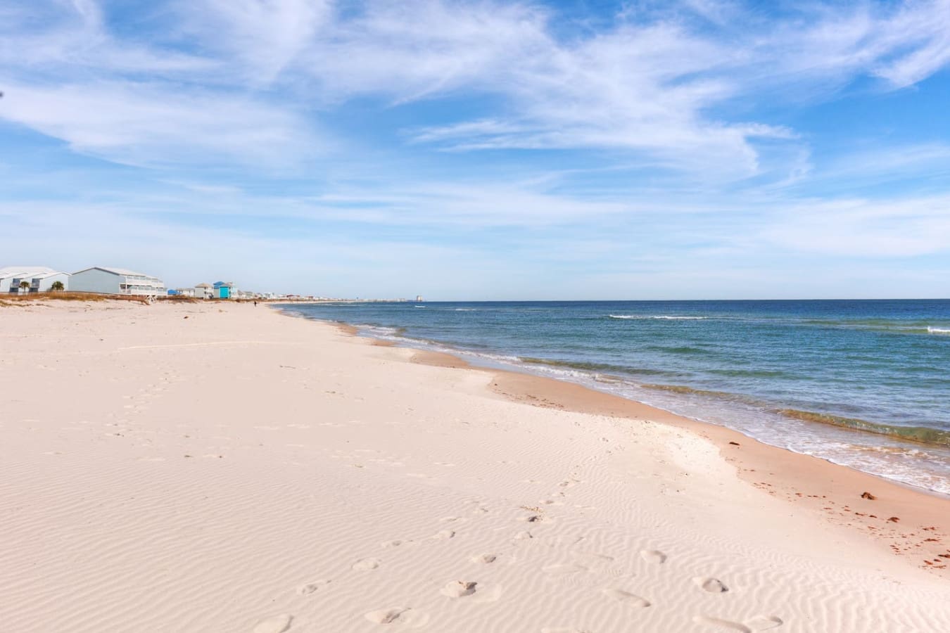 A wide sandy shoreline stretches out peacefully with gentle waves rolling in and a handful of beachfront homes dotting the landscape—this is the kind of quiet, uncrowded beach day most people dream about.