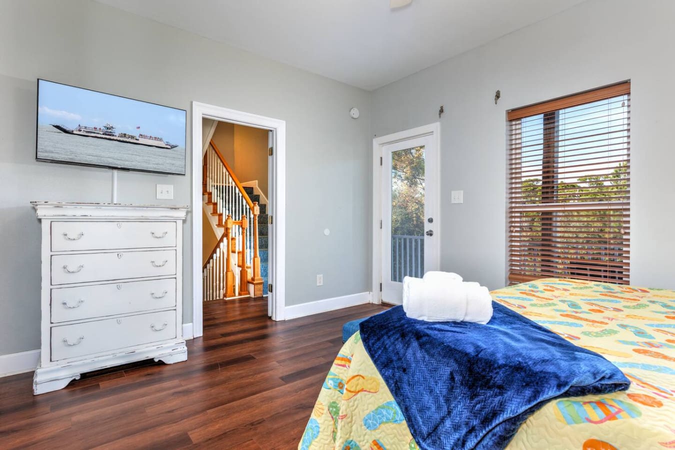 This bright master bedroom nails the coastal vibe with its calming blue walls, cheerful ocean-themed bedding, and those gorgeous wood shutters framing the view while natural light floods in from every angle.