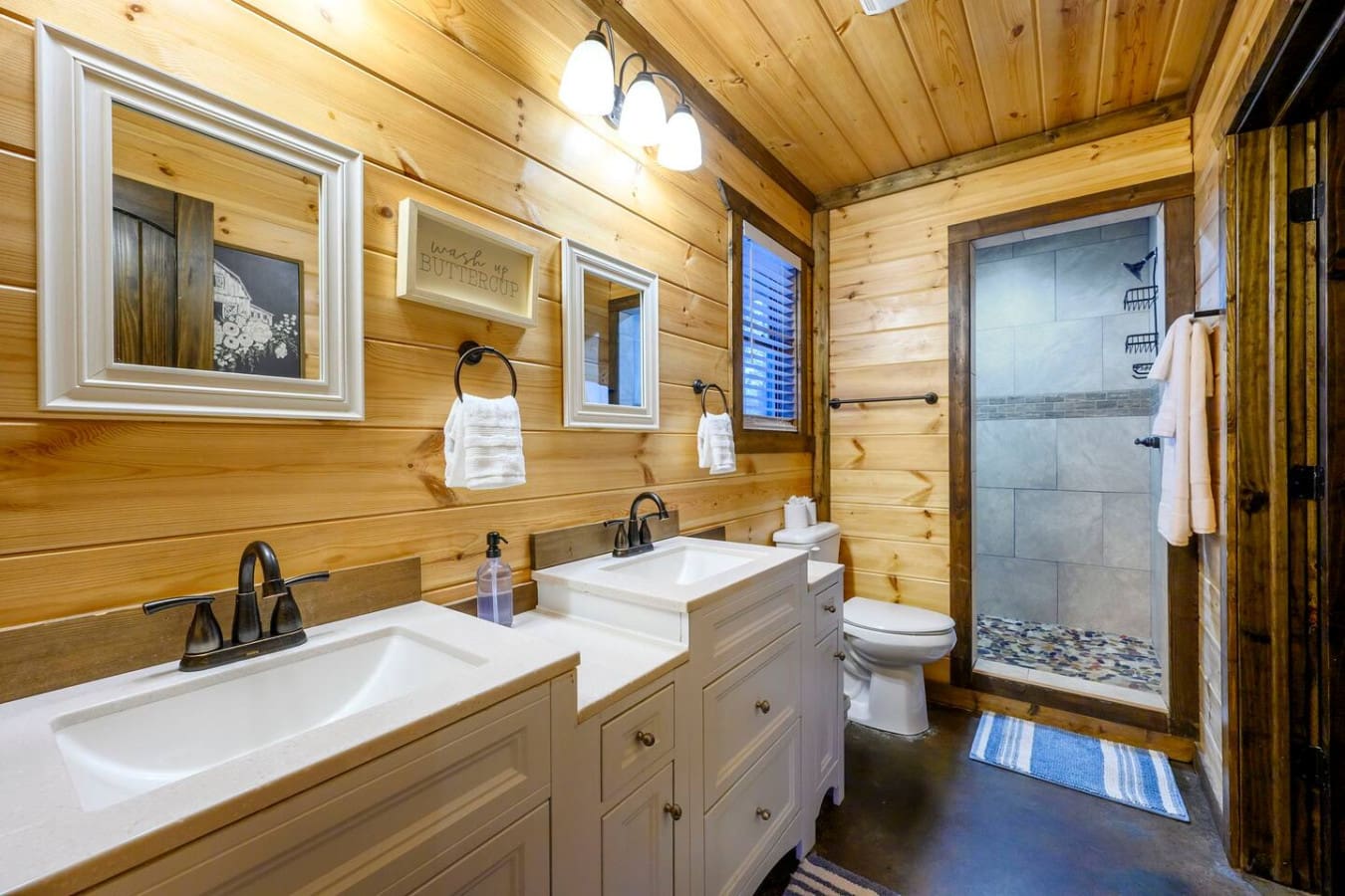 This spacious master bath features double vanities, a walk-in shower, and plenty of natural light in a cozy wood cabin setting.
