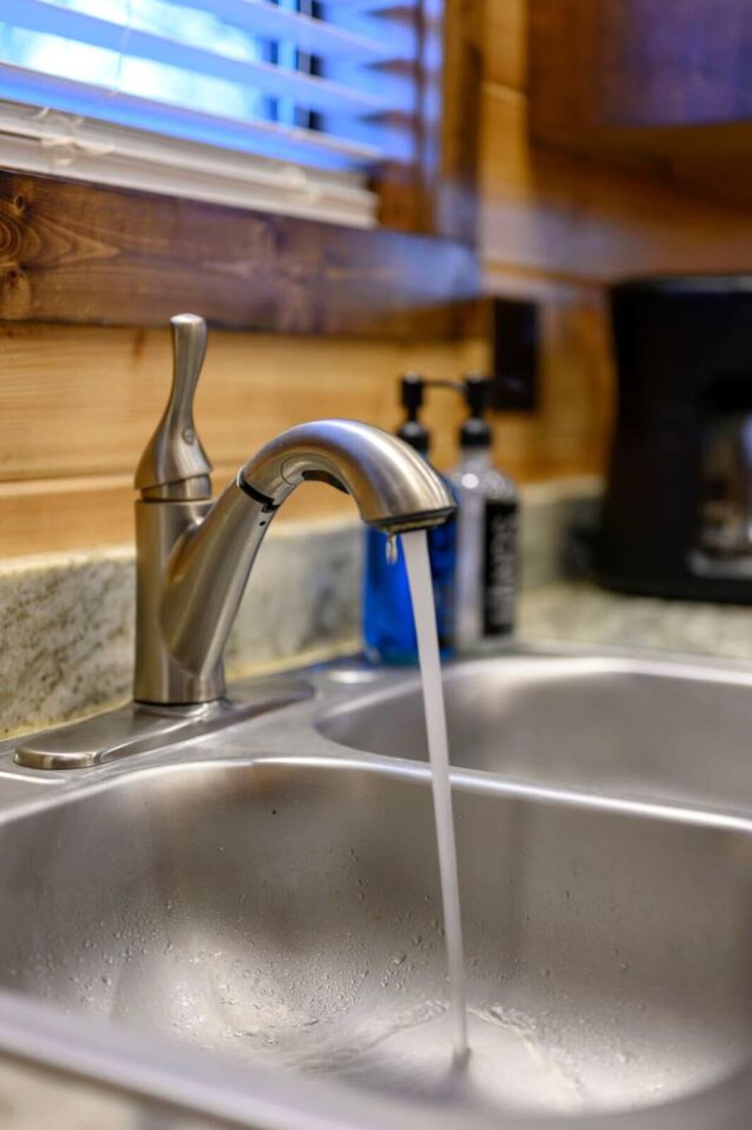 Modern stainless steel faucet flowing into a pristine sink with warm wood cabinetry and ambient blue lighting in the background.
