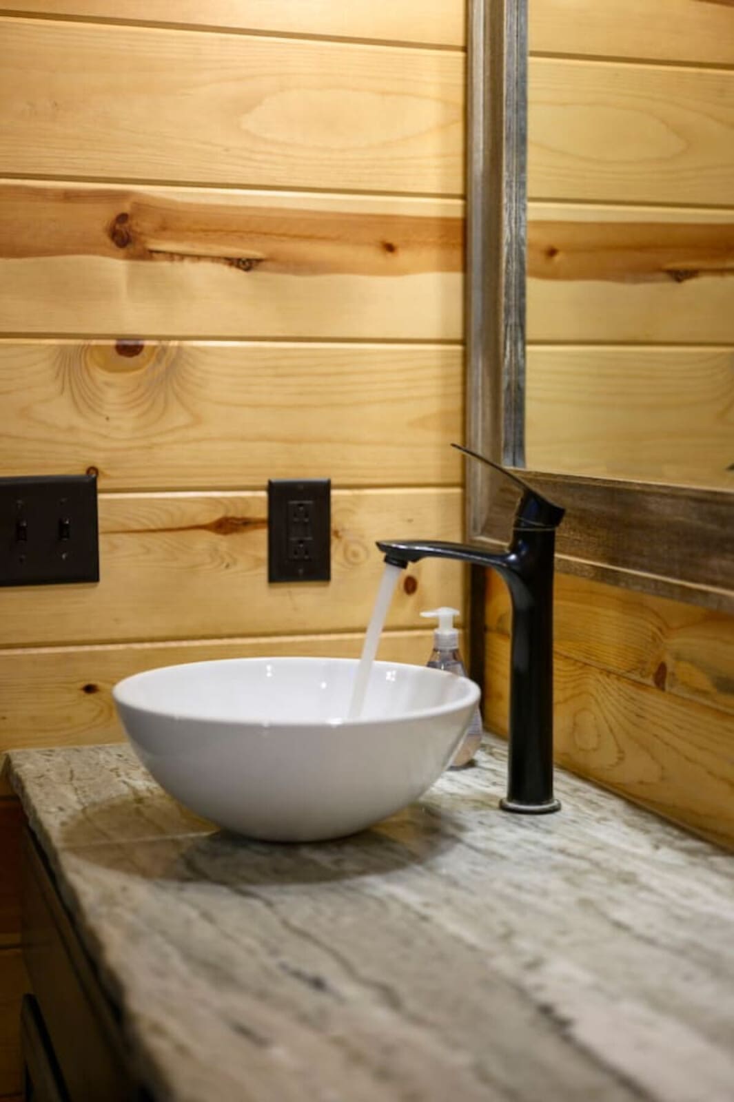 Modern white vessel sink with sleek black faucet sits on marble countertop against rustic wood-paneled walls.
