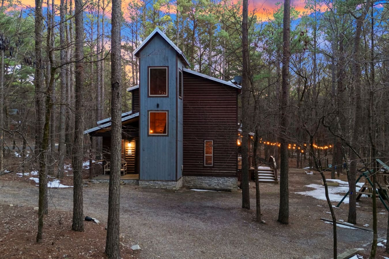 This modern cabin nestled in the woods glows warmly at twilight, surrounded by towering pines and twinkling string lights.