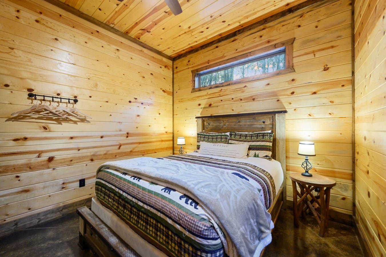 Cozy knotty pine bedroom with a queen bed, warm lighting, and a tranquil forest view through the transom window.
