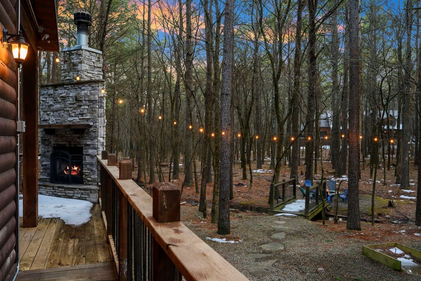 Cozy cabin vibes with an outdoor fireplace overlooking a magical wooded retreat with twinkling lights strung through the trees.