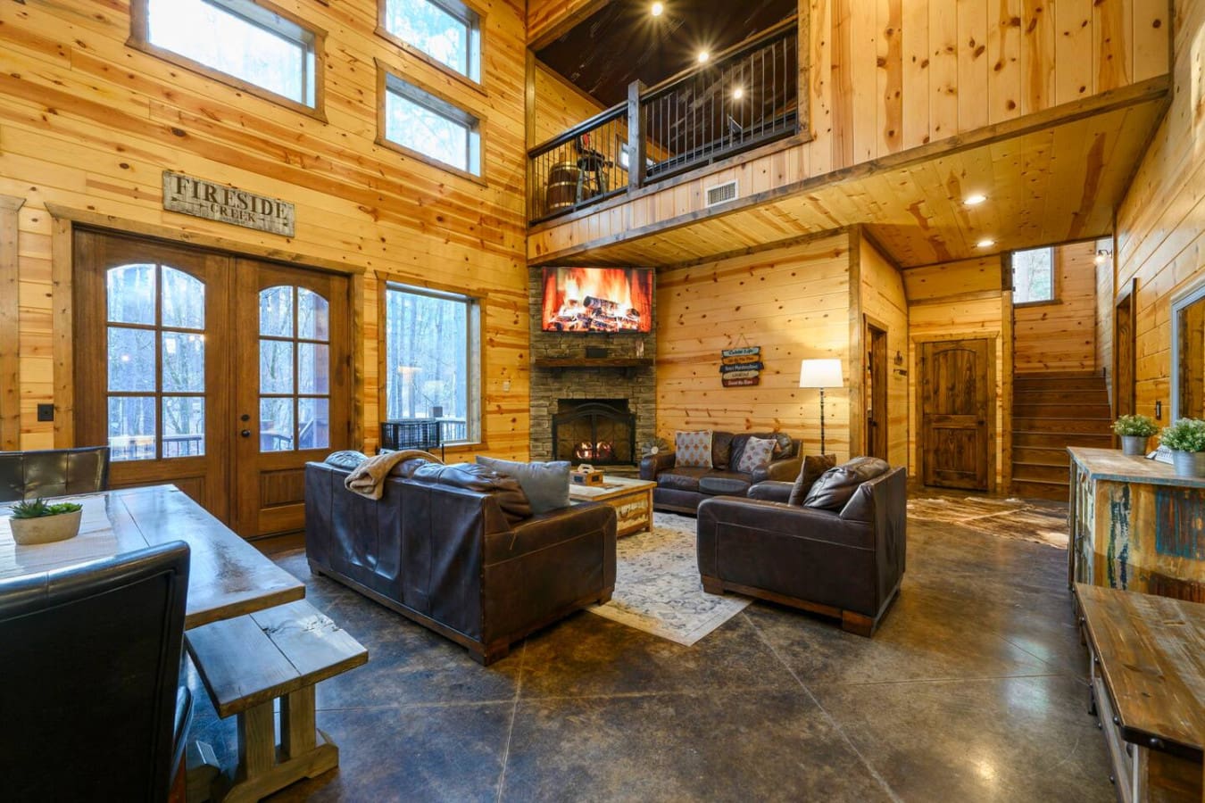 This massive log cabin living room has everything—fireplace, cathedral ceilings, a loft, and those huge windows looking out at the trees.