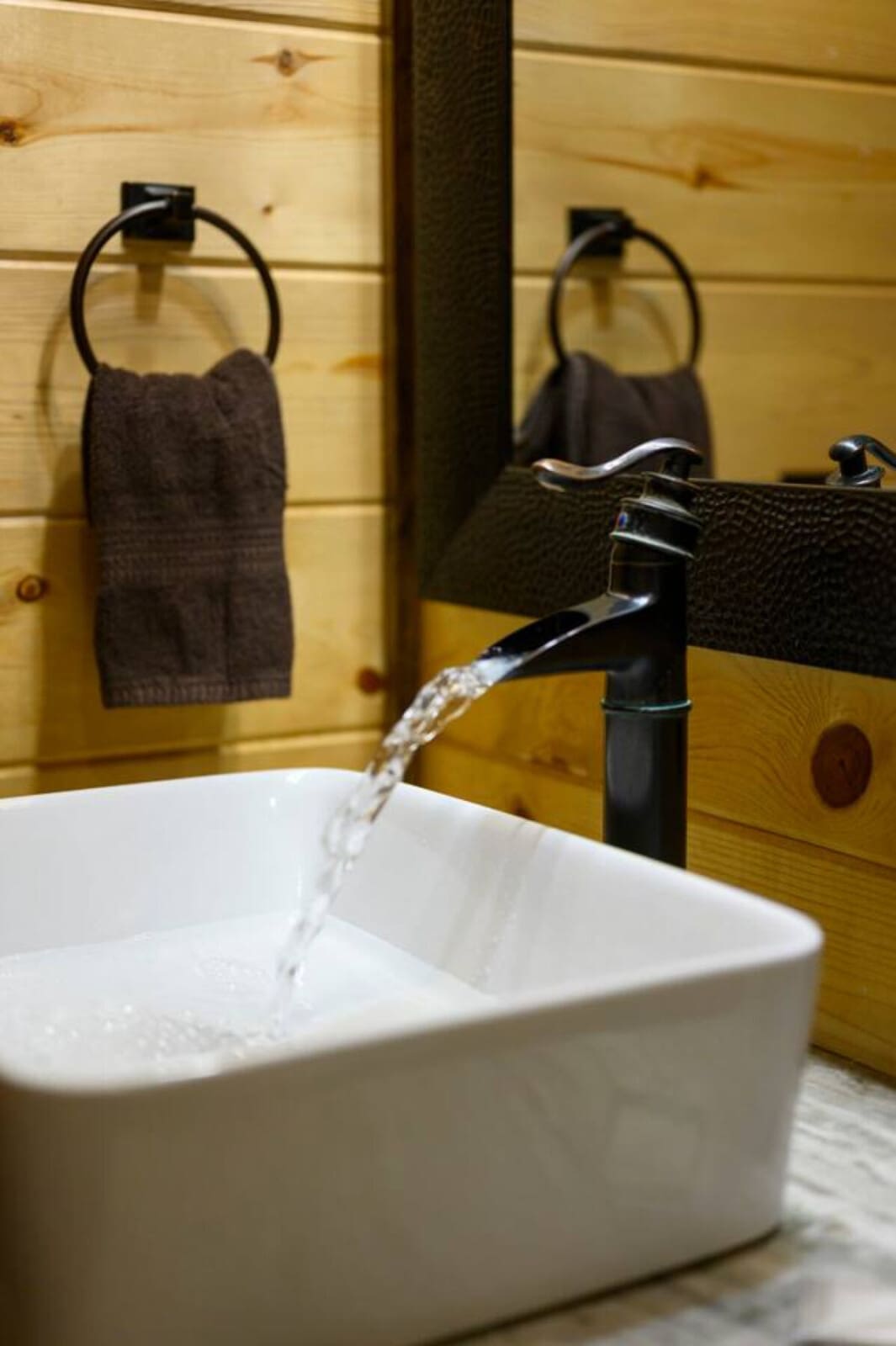 Water flows from a sleek black faucet into a white vessel sink against natural wood walls.