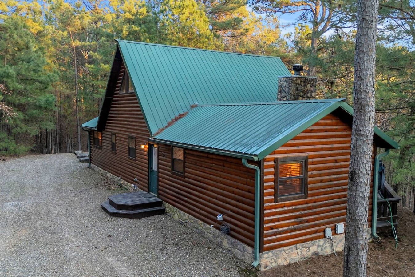 This cozy log cabin sits nestled in the forest with a striking turquoise metal roof and stone foundation.