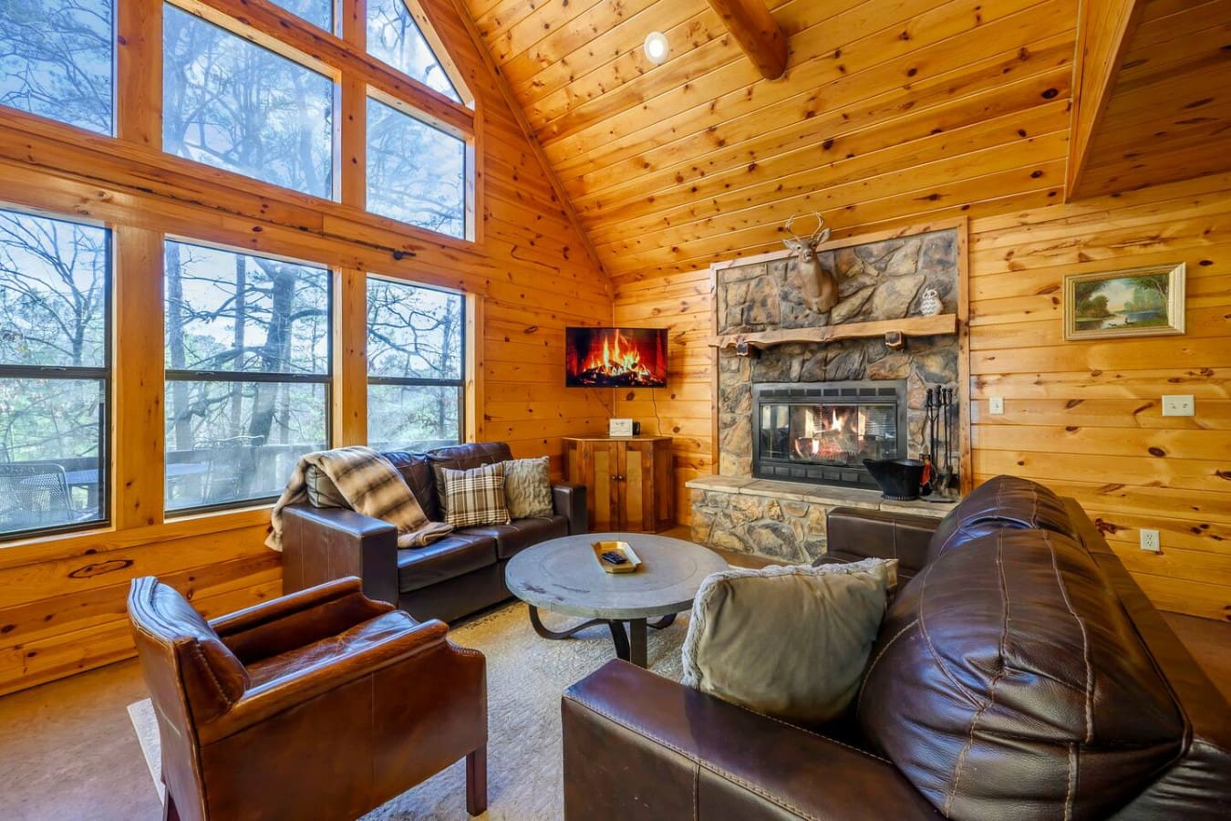 This cozy log cabin living room features a stone fireplace, leather seating, and floor-to-ceiling windows overlooking the forest.