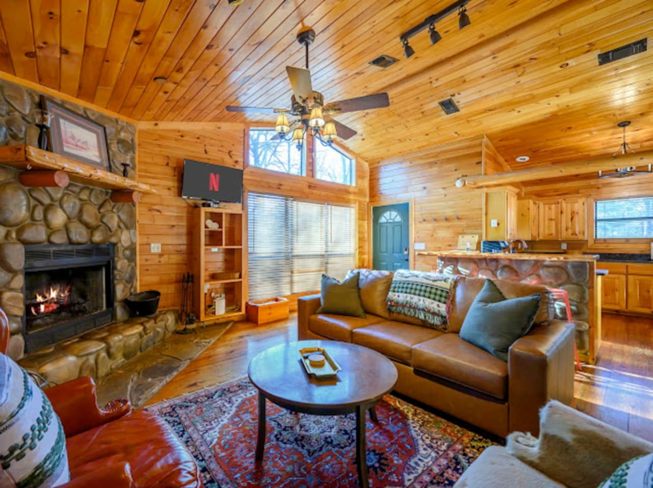 This cozy log cabin living room features a stone fireplace, vaulted wood ceilings, and plenty of natural light for a perfect retreat.