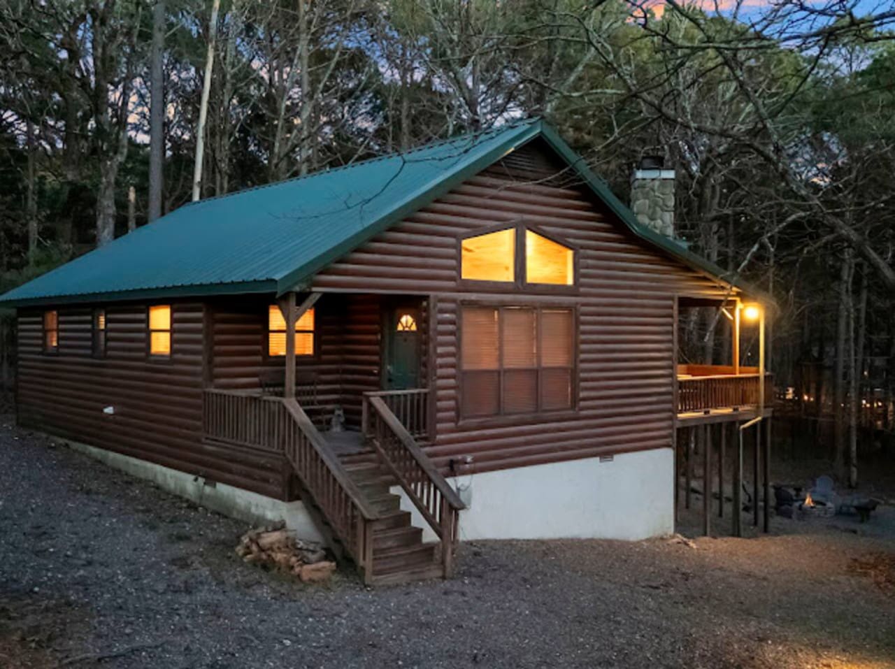Cozy log cabin nestled in the woods with a wraparound deck and warm interior lighting at dusk.