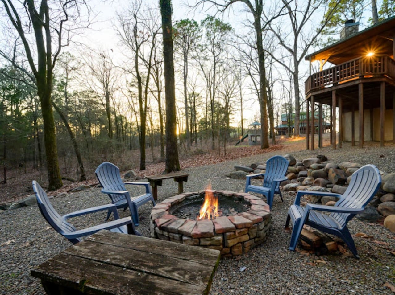 Cozy fire pit surrounded by tall pines with blue Adirondack chairs and a modern elevated deck in the background.