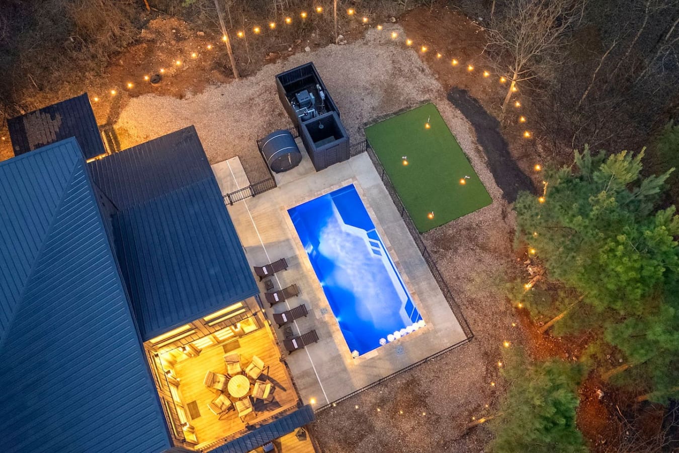 Aerial view of your private backyard oasis with glowing pool, putting green, and cozy fire pit surrounded by twinkling lights.