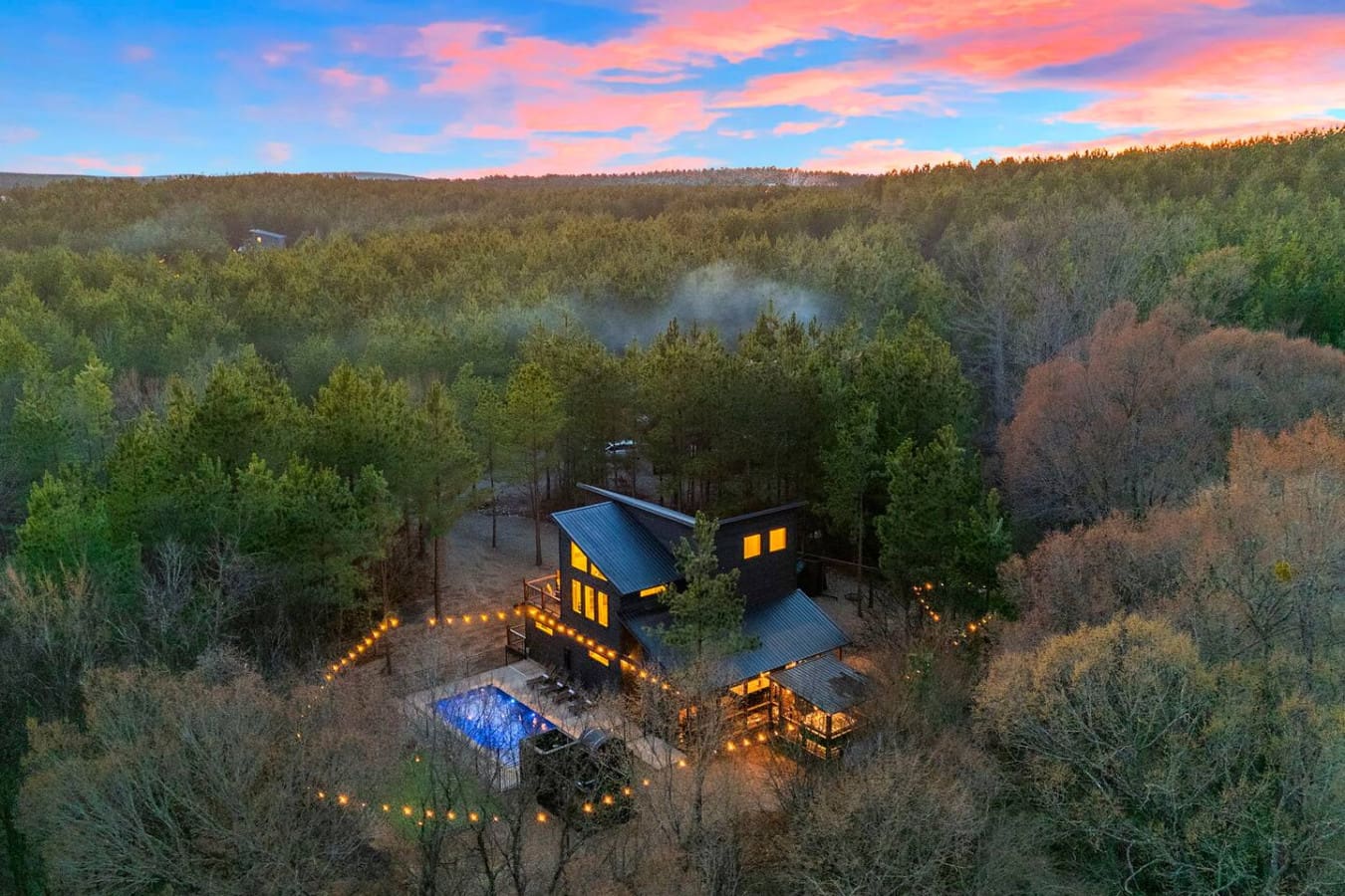 This stunning modern cabin sits nestled in the forest with a glowing pool and pathways, surrounded by towering trees and mountain views.