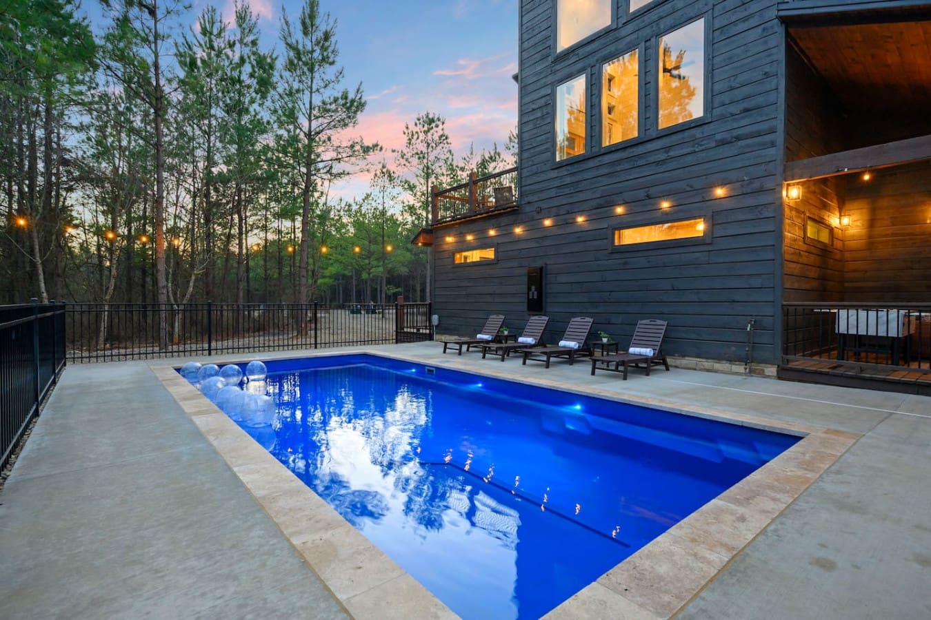 This stunning modern home features an illuminated resort-style pool with lounge seating overlooking a serene forested backdrop.