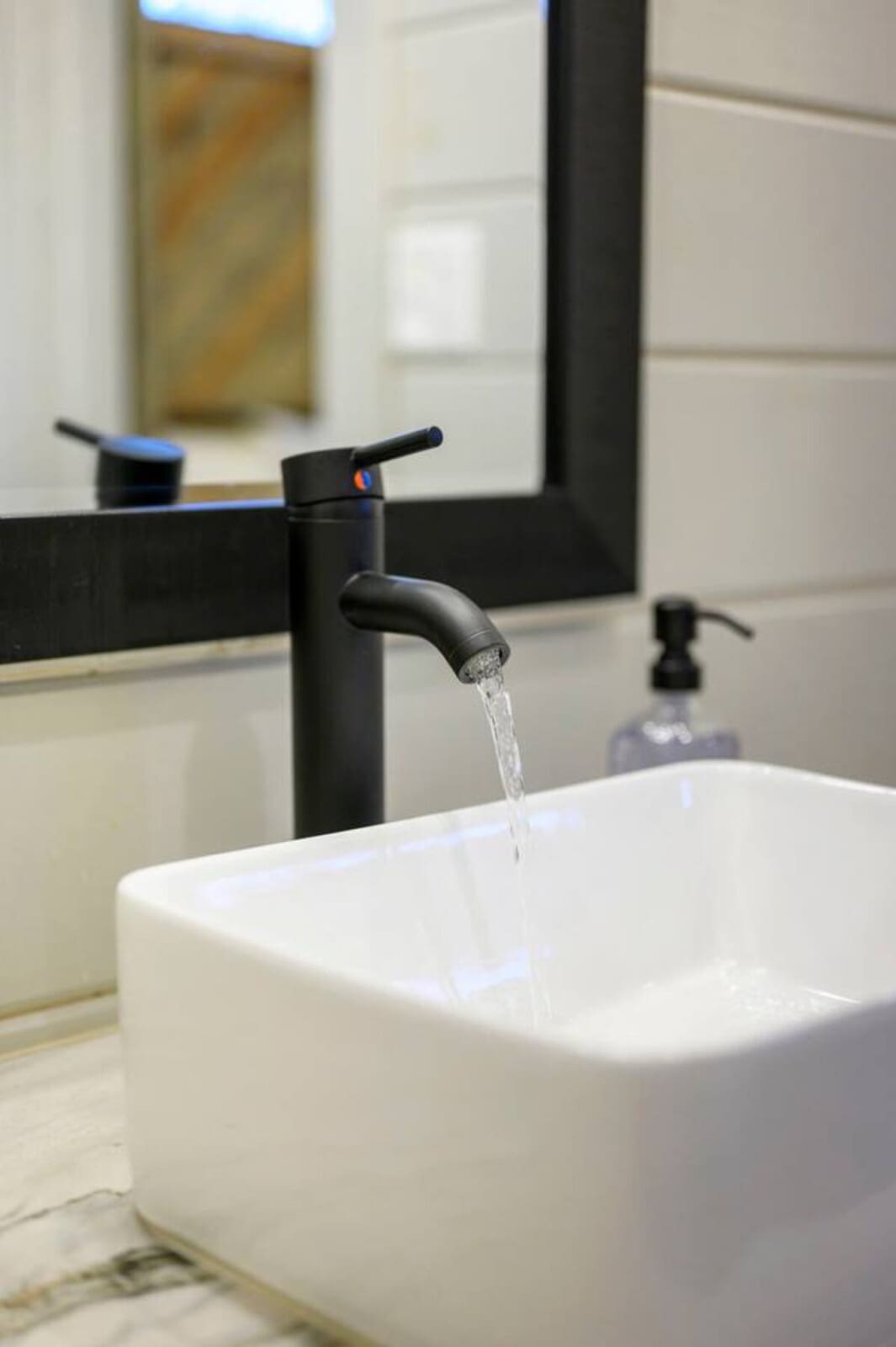 Modern black faucet flowing water into a sleek white vessel sink with a mirror and spa-like bathroom behind it.