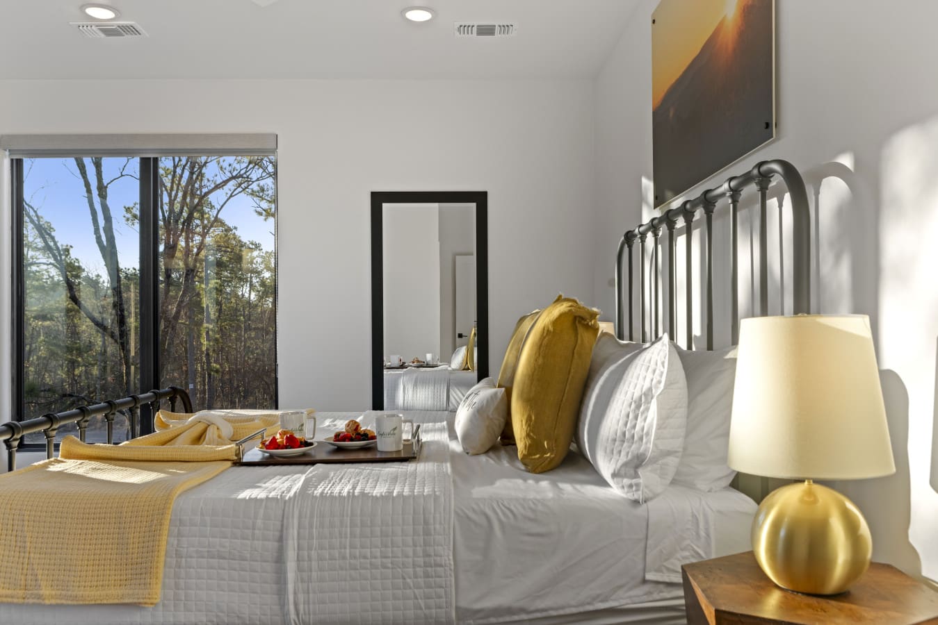 This bright master bedroom features a stunning forest view, a sleek metal bed frame, and warm gold accents throughout.