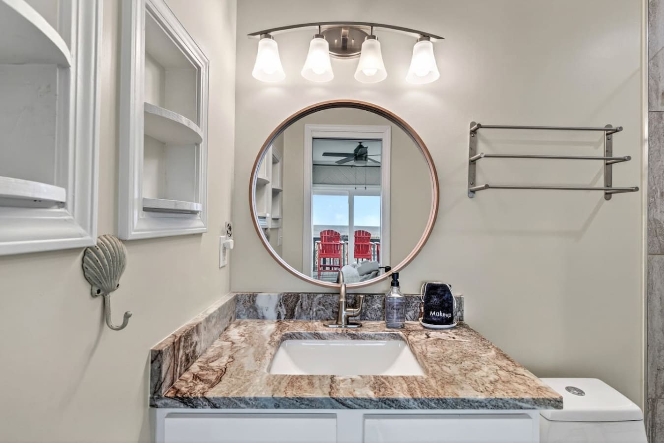This stunning bathroom features a gorgeous marble vanity, sleek fixtures, and a circular mirror framing an amazing view of the waterfront and Adirondack chairs.