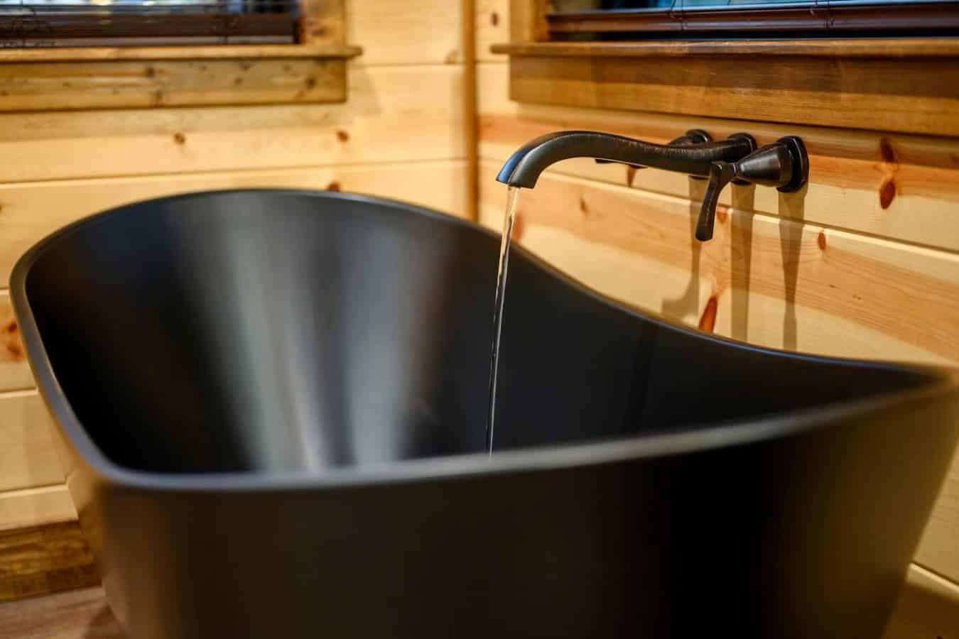 Black soaking tub with wall-mounted faucet overlooking the wooden cabin interior and windows.