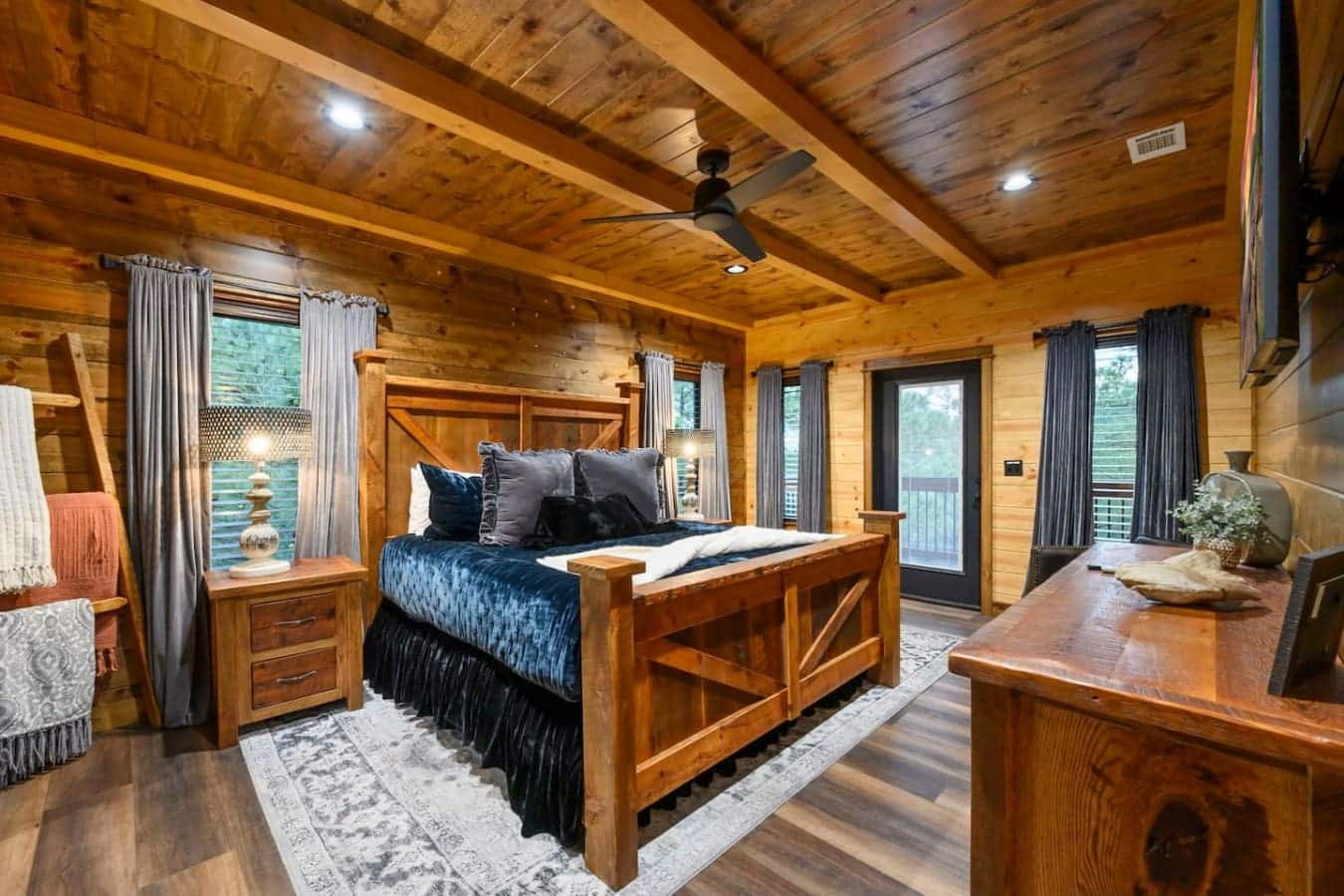 This luxurious log cabin bedroom features exposed wood beams, a custom wooden bed frame, and multiple glass doors opening onto forest views.