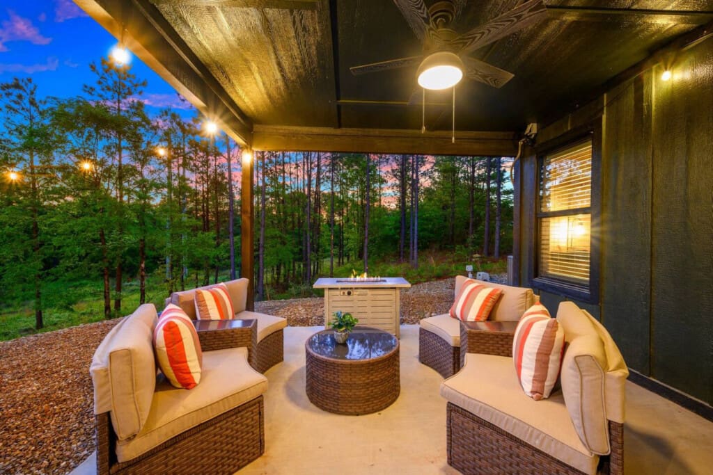 As the sun dips below the treeline, this covered patio becomes your own little forest sanctuary—surrounded by cozy seating, a glowing fire pit, and those towering pines creating the perfect backdrop for the evening ahead.