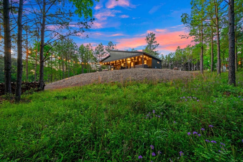 Watch this sleek modern cabin come alive as the sun sets behind the pines, casting that perfect golden glow across the gravel courtyard while the sky turns those dreamy shades of pink and blue.