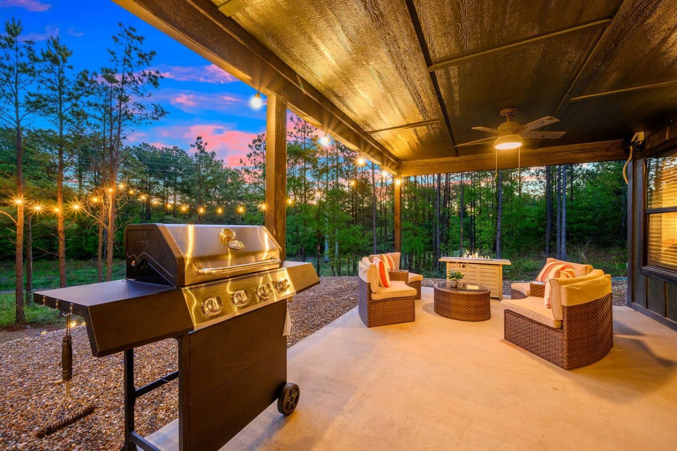 This outdoor setup is basically made for hanging out all evening—a killer grill ready to go, cozy seating scattered around, warm overhead lighting, and that forest view getting better as the sky turns those gorgeous twilight colors.
