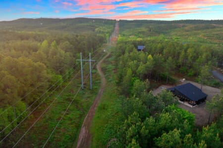Your own slice of forest is waiting with power lines stretching into the distance, a modern cabin glowing in the clearing, and nothing but endless green pines rolling out in every direction as the sky turns that perfect sunset pink.