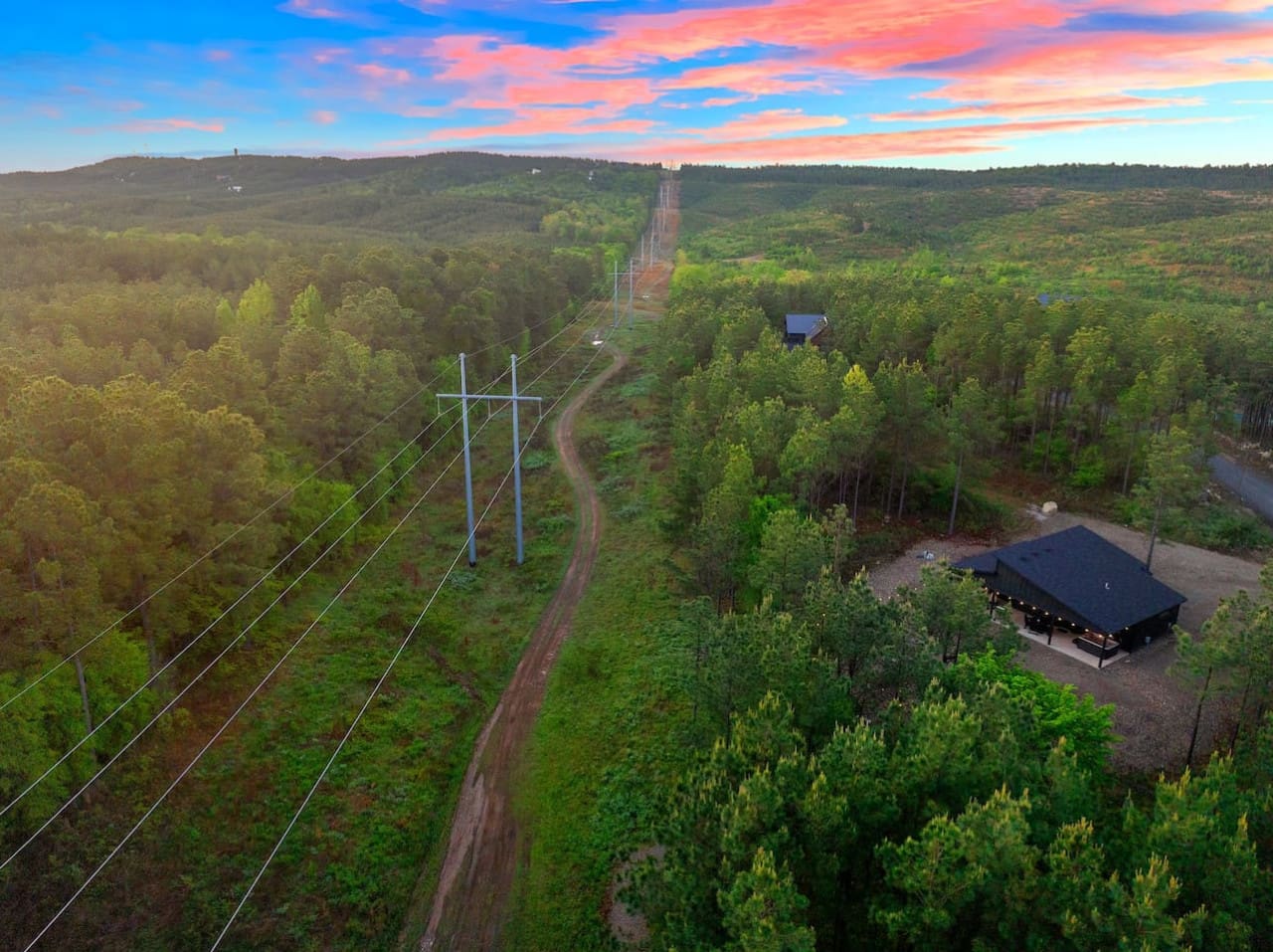 Your own slice of forest is waiting with power lines stretching into the distance, a modern cabin glowing in the clearing, and nothing but endless green pines rolling out in every direction as the sky turns that perfect sunset pink.