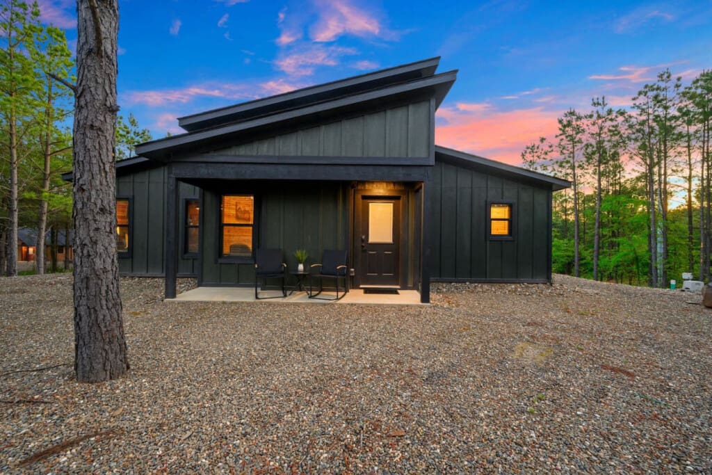This sleek modern black cabin tucked into the forest feels like your own private sanctuary, with warm light spilling onto the gravel courtyard as the sky turns that perfect twilight blue.