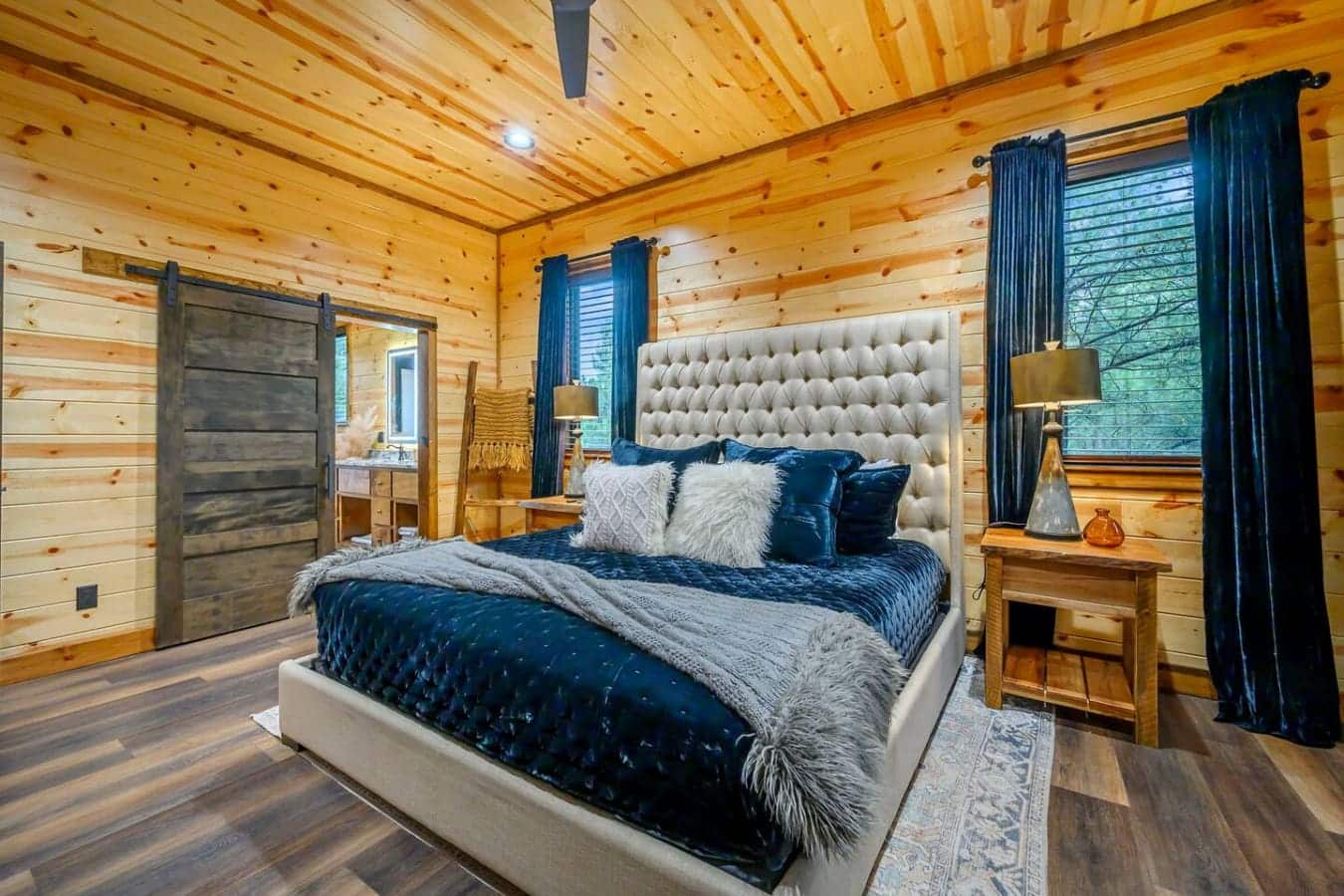 This stunning log cabin bedroom features a plush tufted headboard, cozy wood walls, and elegant teal curtains framing the forest views.