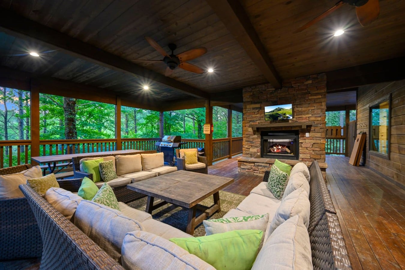 Outdoor living area with fireplace, TV, sectional seating, and forest views.